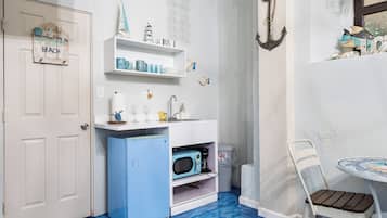 A room with a blue floor, white walls, a white door, a small kitchenette, a white chair, and a hanging light fixture.
