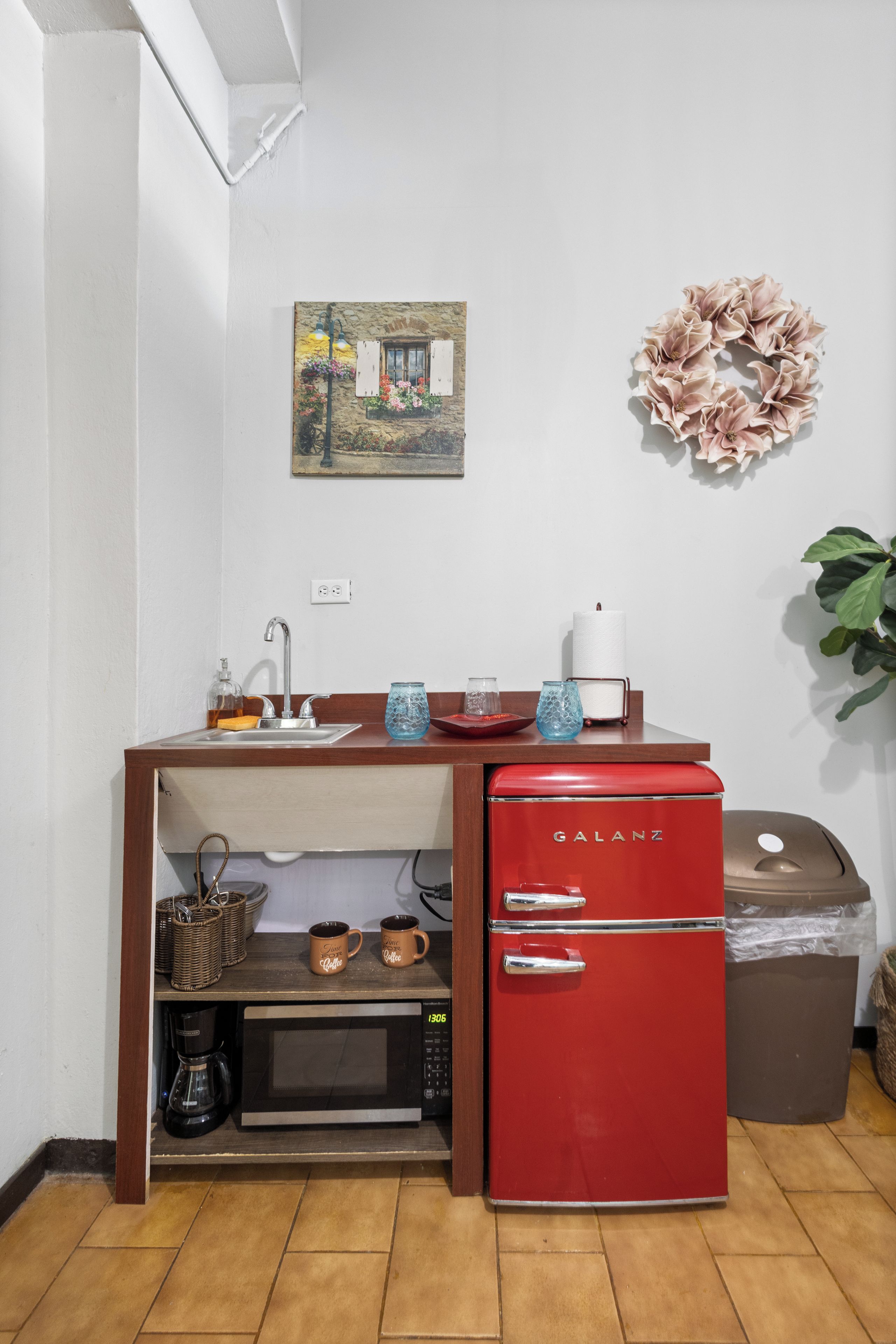 A compact kitchenette with a red Galanz refrigerator, a microwave, a sink, and a small shelf with a coffee maker and mugs.