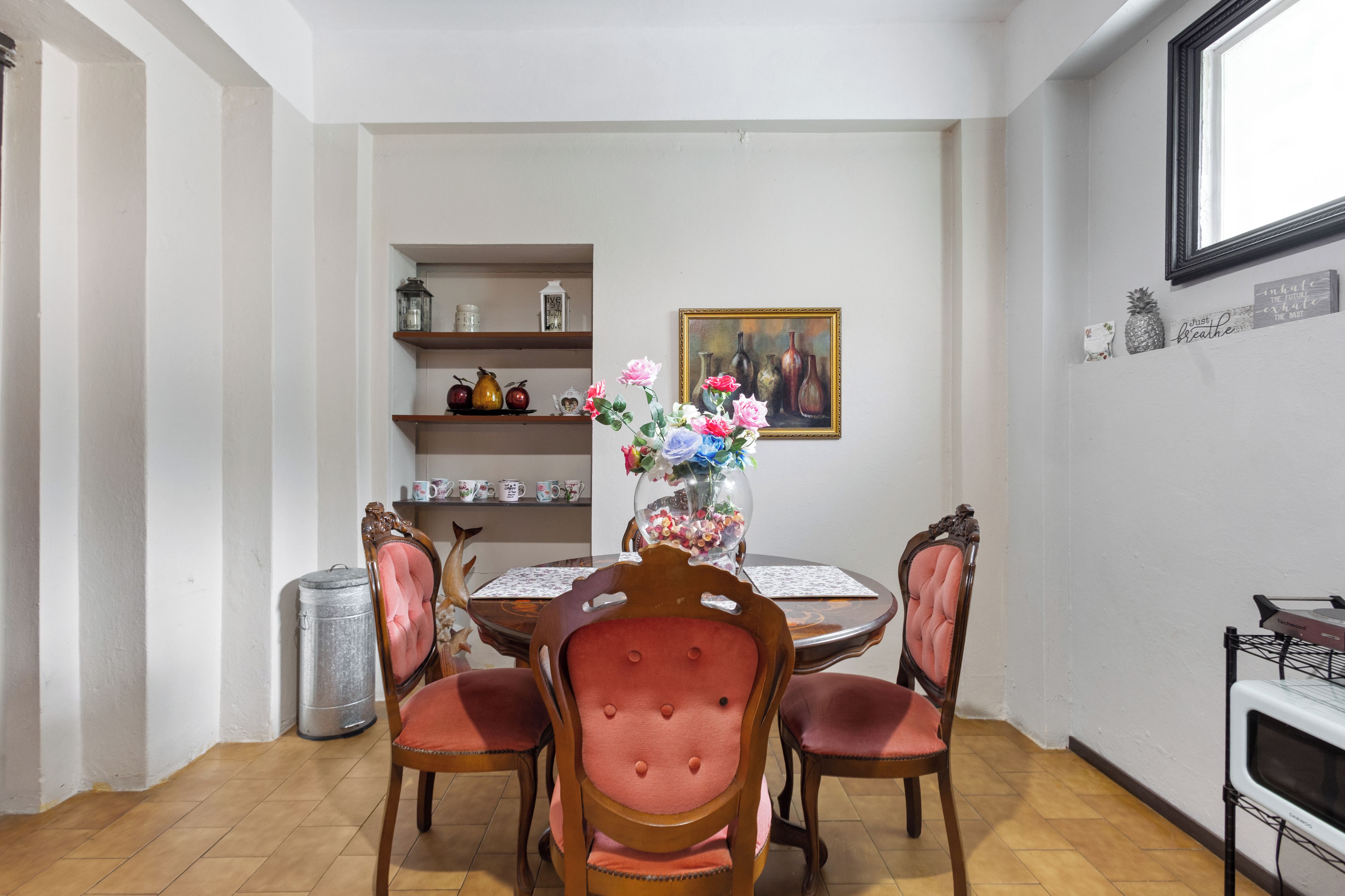 A dining area with a round table, four chairs, a vase with flowers, a painting, and shelves with decorative items.