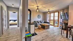 A spacious living area with a sofa, coffee table, and a fireplace. There is a dining table with chairs and a TV. The room has wooden flooring and large windows. - Apartamenty Sun & Snow Karpacz Myśliwska (Karpacz)