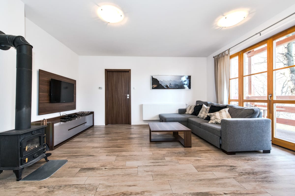 A modern living room with a wood-burning stove, a grey sofa, a flat-screen TV, and a large window with a view.