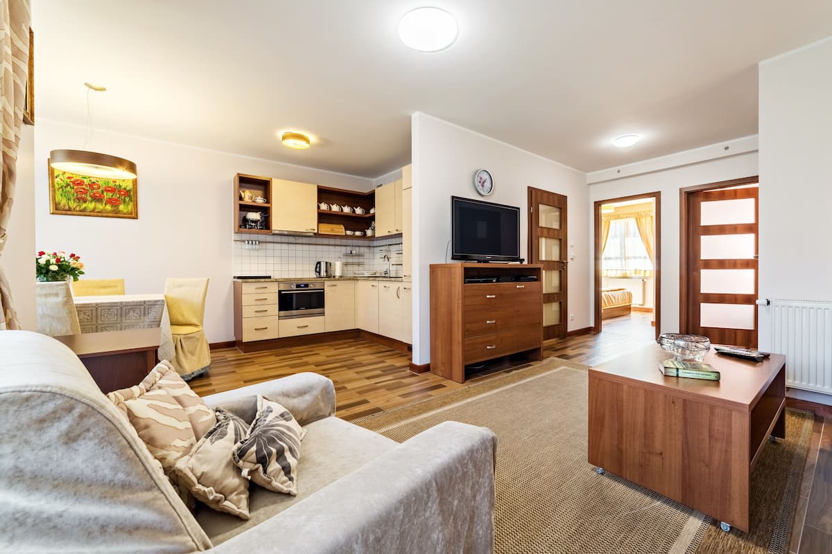 A modern living room with a sofa, a coffee table, and a TV stand. There is a kitchen area with cabinets and a dining table.