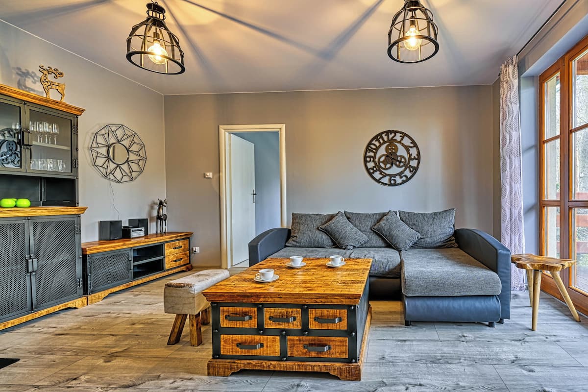 A cozy living room with a sofa, a wooden coffee table, and a dining area with a cabinet. There are two wall clocks and a window with curtains.
