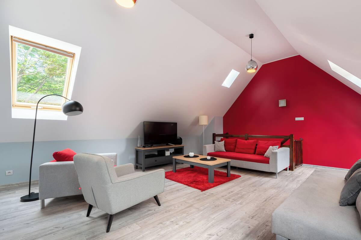 A modern loft apartment with a sloped ceiling, a red accent wall, a large window, and a flat-screen TV.