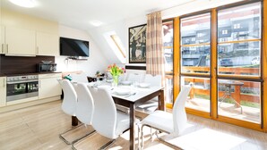 A modern kitchen with a flat-screen TV, a dining area with white chairs and a table set for four, and a large sliding glass door leading to a balcony with a view of buildings and a car. - Apartamenty Sun & Snow Karpacz Myśliwska (Karpacz)