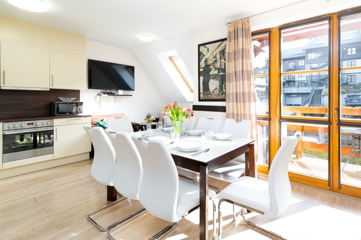 A modern kitchen with a flat-screen TV, a dining area with white chairs and a table set for four, and a large sliding glass door leading to a balcony with a view of buildings and a car.