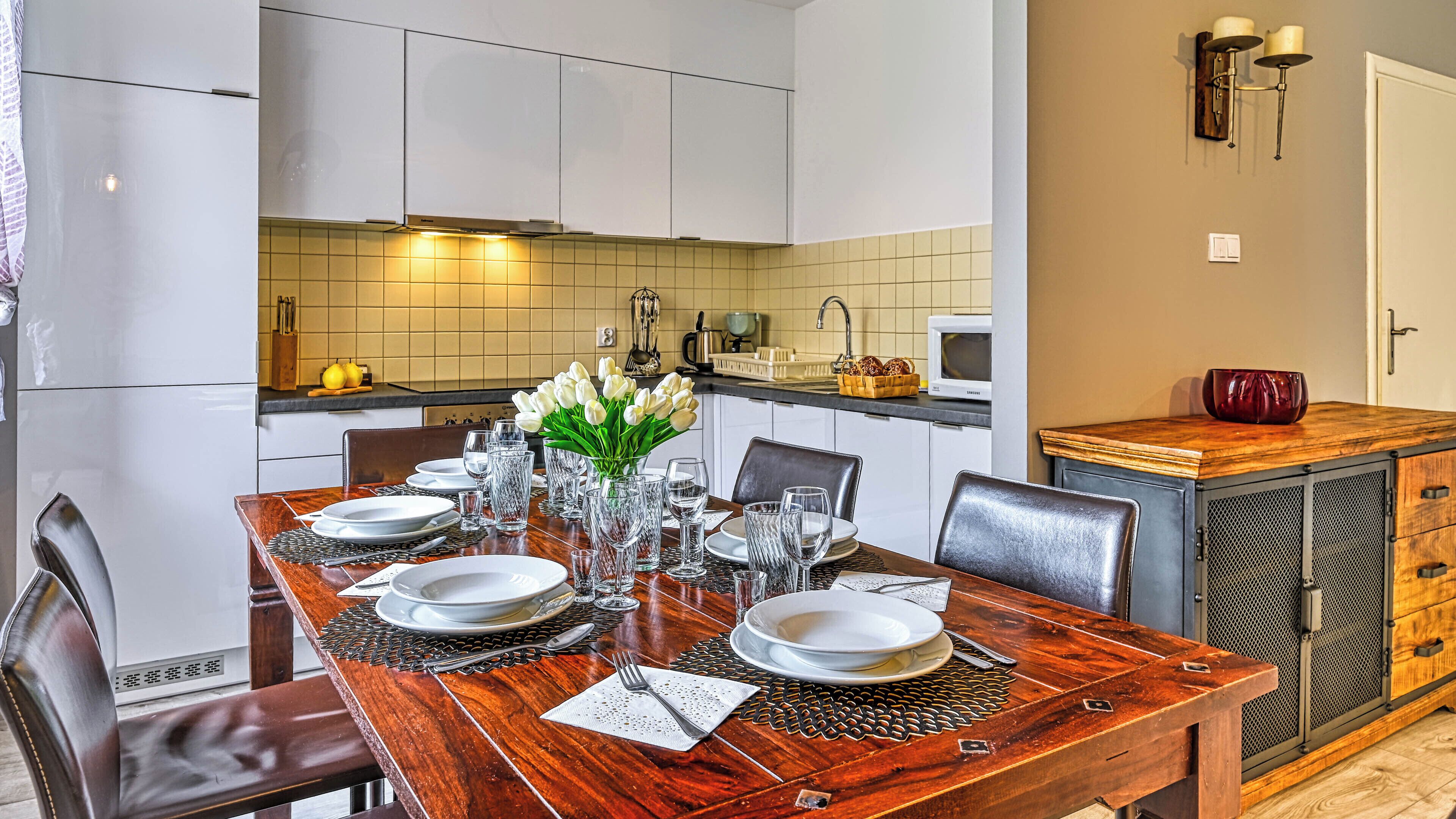 Un coin repas avec une table en bois, dressée pour quatre personnes, entourée de chaises. Un vase contenant des fleurs blanches est placé au centre.