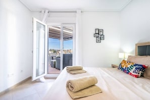 A hotel room with a bed, a nightstand, a lamp, a window with a view of the city, and a door leading to an outdoor area.