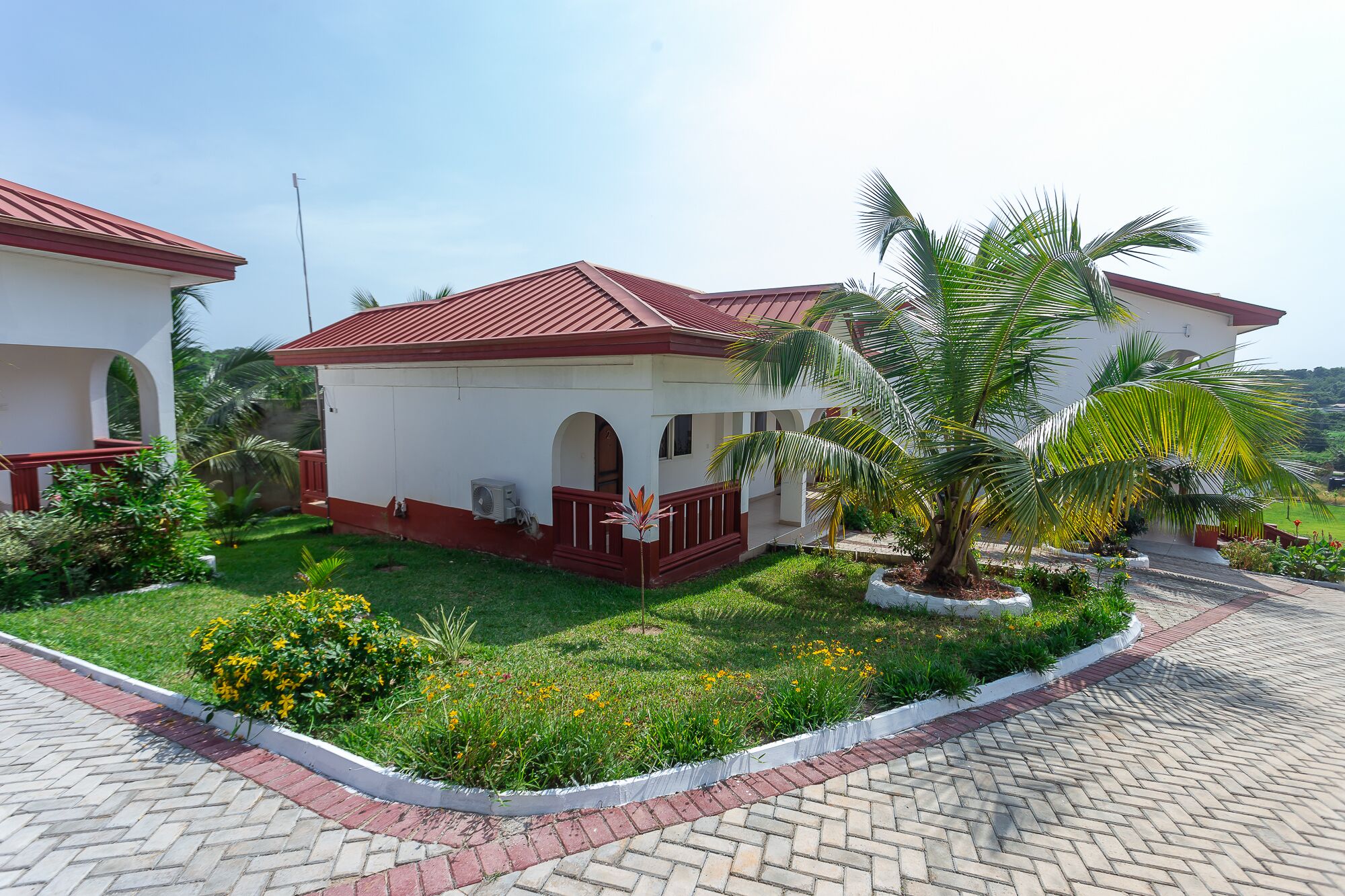 A two-story building with a red roof and white walls, surrounded by a well-maintained garden with palm trees and flowering plants.