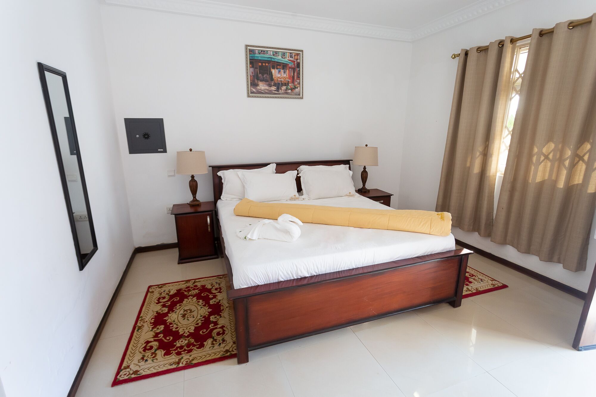 Una cama impecablemente arreglada con sábanas blancas y una manta amarilla, un cabecero de madera, dos mesitas de noche con lámparas, una alfombra con estampado rojo y dorado, un cuadro en la pared y una ventana con cortinas color beige.