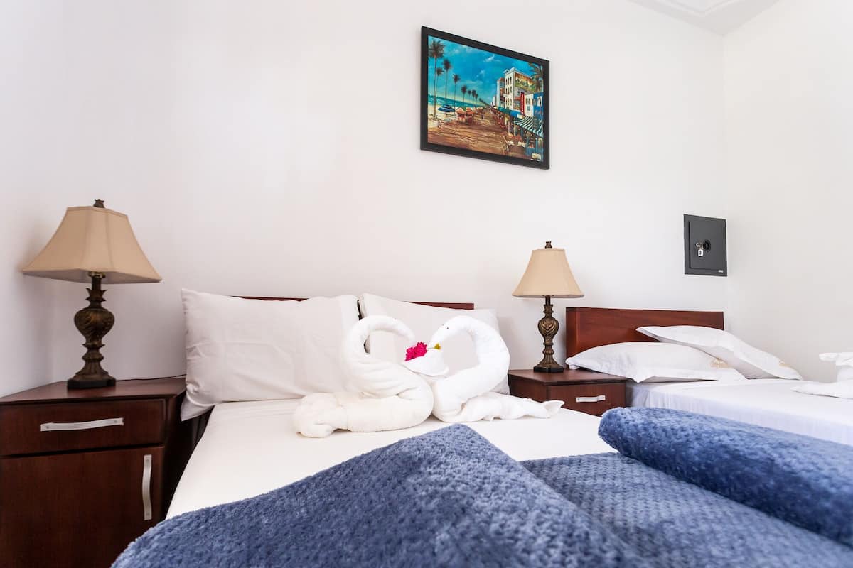 A hotel room with a bed, two bedside tables, a lamp, a safe, and a painting on the wall.