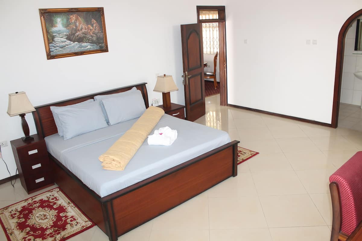 A neatly made bed with a blue mattress and white pillows, a wooden headboard, a bedside table with a lamp, and a red patterned rug on the floor.