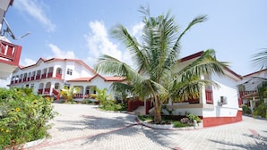 A courtyard with a palm tree, a red and white building, and a paved walkway. - Royal Elmount Hotel (Elmina)