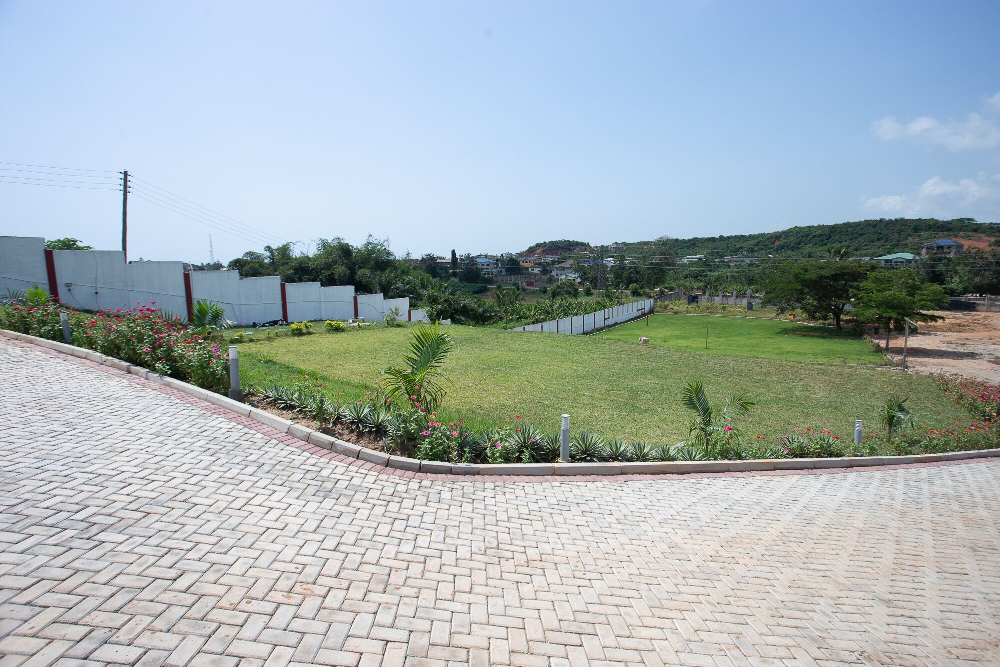 A paved area with a grassy field, a white fence, and a hilly landscape in the background.