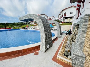 A swimming pool with stone arches, outdoor seating, and a view of nearby buildings. - Royal Elmount Hotel (Elmina)