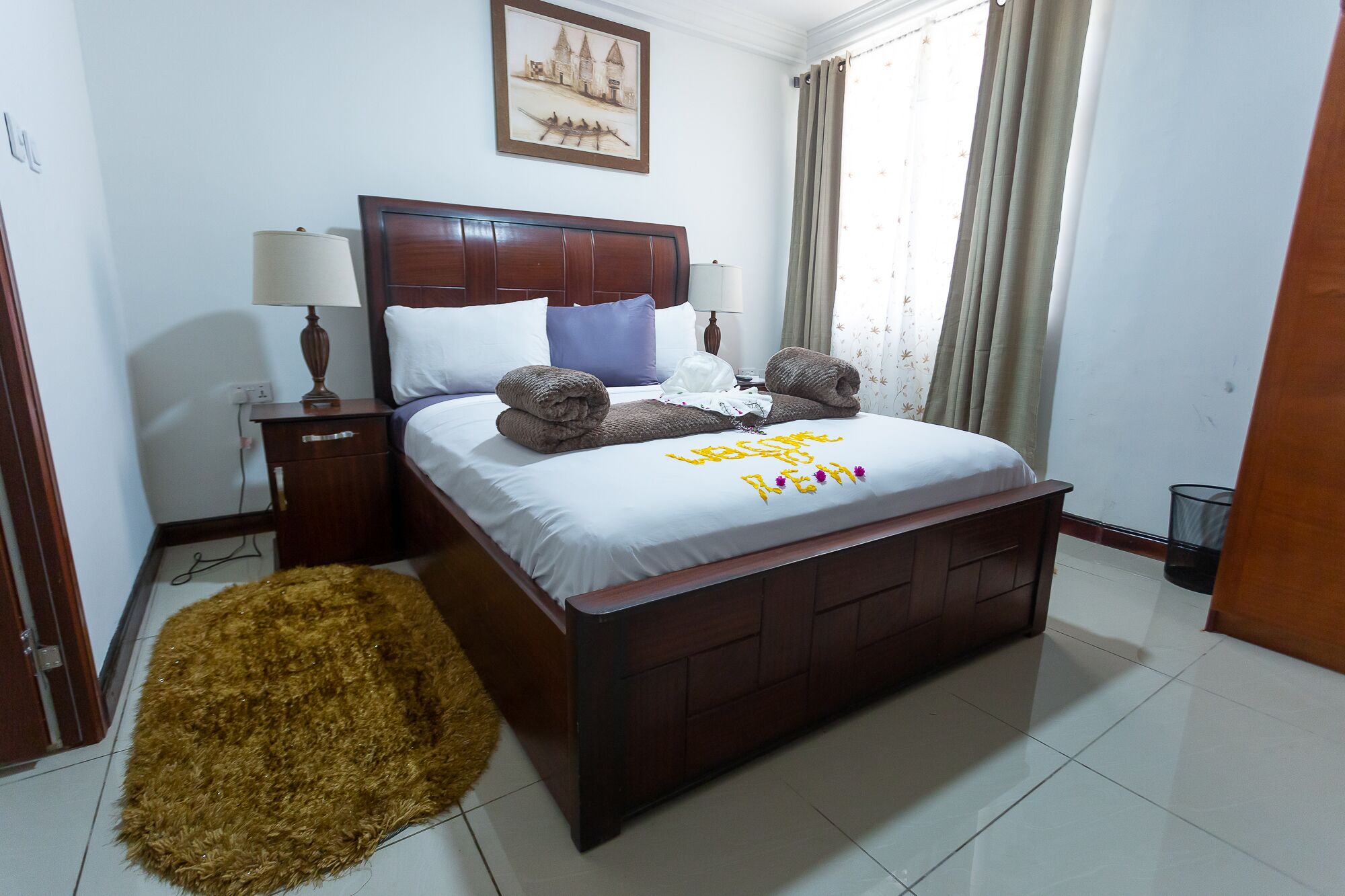 A neatly made bed with a white comforter and decorative pillows, a wooden headboard, a bedside table with lamps, and a framed picture on the wall.