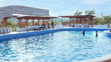 A pool area with lounge chairs and umbrellas, surrounded by a wooden pergola and a view of greenery.