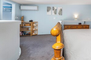 A bedroom with a bed, a wooden headboard, a dresser, a shelf with books and decorative items, and a wall-mounted air conditioner.
