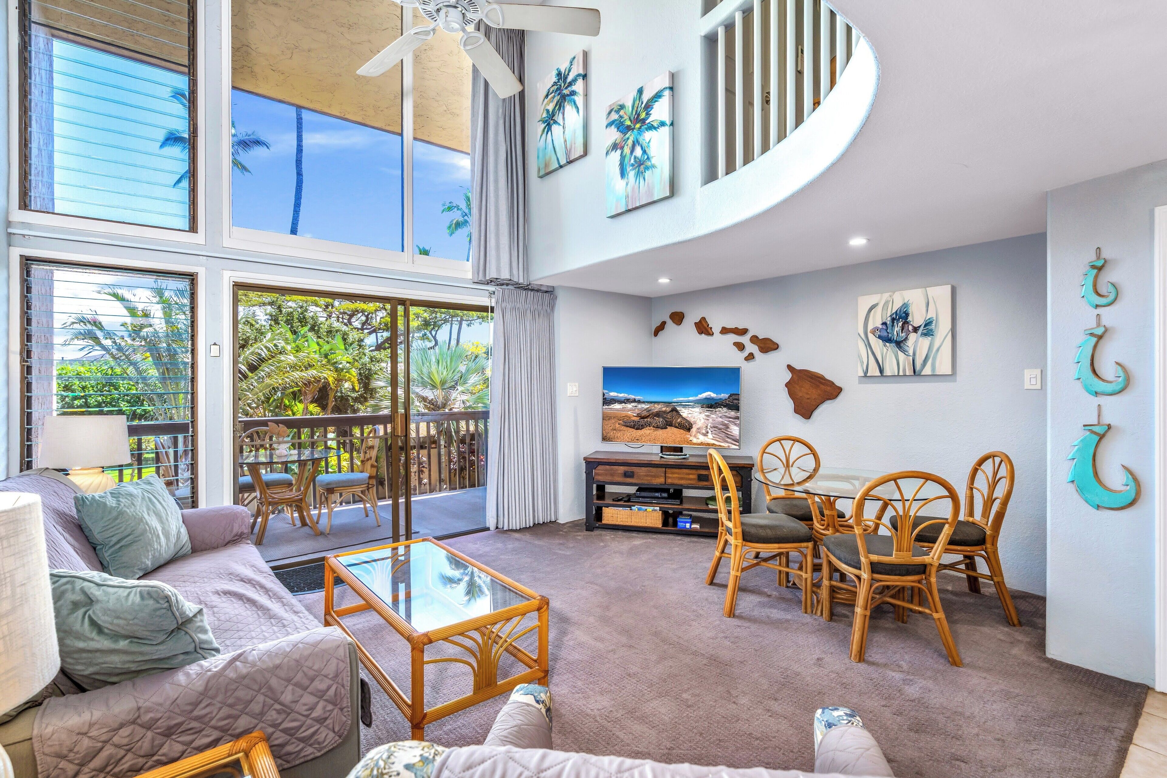 A spacious living room with a large window, a ceiling fan, a television, and a dining area with wicker furniture.