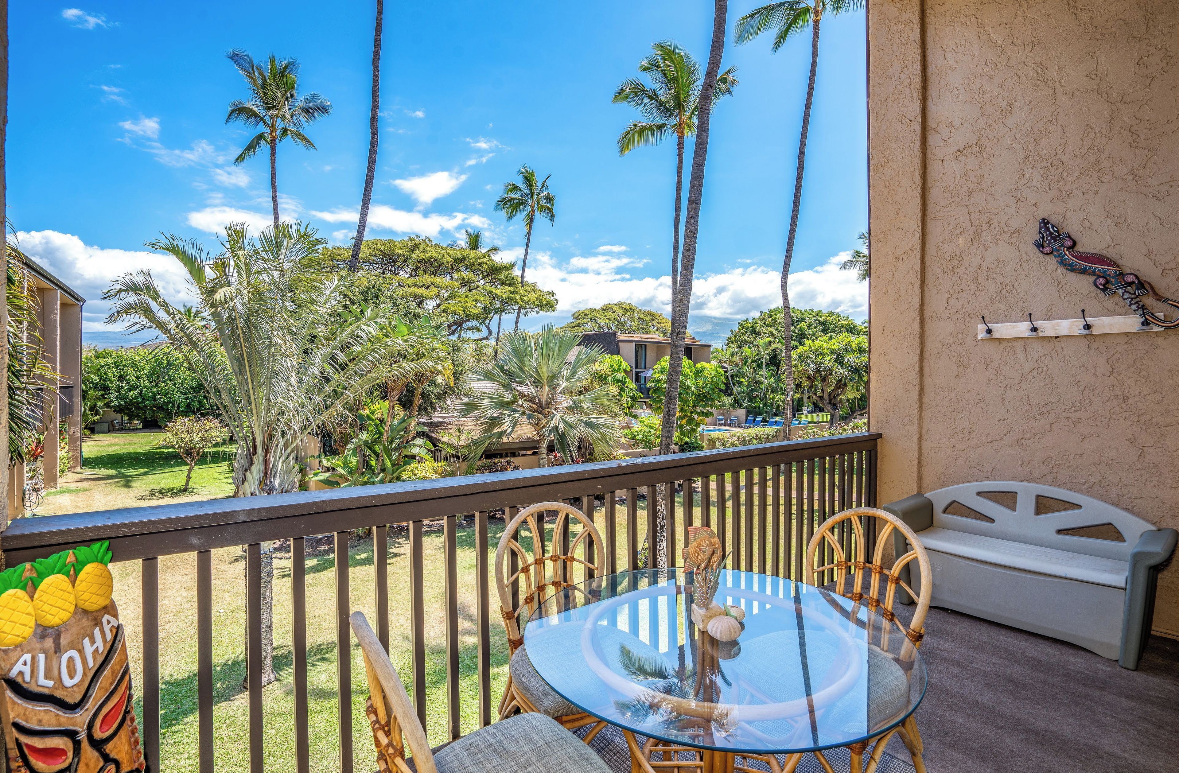 A glass table with wicker chairs on a balcony overlooking a garden with palm trees and a view of a building.