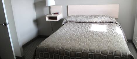Habitación de hotel con cama, mesita de noche con lámpara, estante en la pared y perchero.