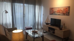 Ett vardagsrum med ett soffbord i glas, en soffa, en tv-apparat placerad på ett träskåp, en lampa och en tavla som föreställer en stadsbild.