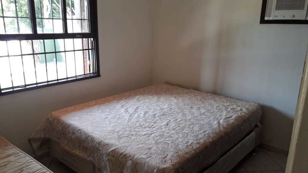 Ein kleines Zimmer mit einem Einzelbett, einer Bettdecke mit Muster, einem Fenster mit Gitter und einer schlichten Wand.