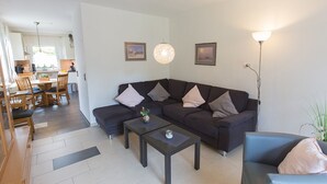 A modern living room with a dark sofa, a coffee table, and a dining area with wooden furniture. - Vacation home Lueg, holiday apartment rental North Sea - Holiday home Lueg (Varel)