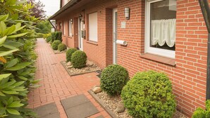 A row of brick buildings with a paved walkway, each with a door and window, and surrounded by well-maintained shrubs. - Vacation home Lueg, holiday apartment rental North Sea - Holiday home Lueg (Varel)