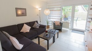A modern living room with a sofa, armchairs, a coffee table, a floor lamp, and a sliding glass door leading to an outdoor area. - Vacation home Lueg, holiday apartment rental North Sea - Holiday home Lueg (Varel)