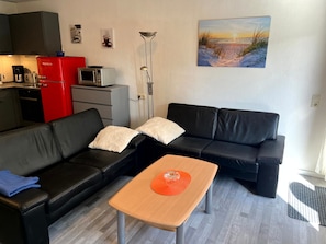 A modern living room with a black leather sofa, a wooden coffee table, and a kitchen area with a red refrigerator and a microwave.