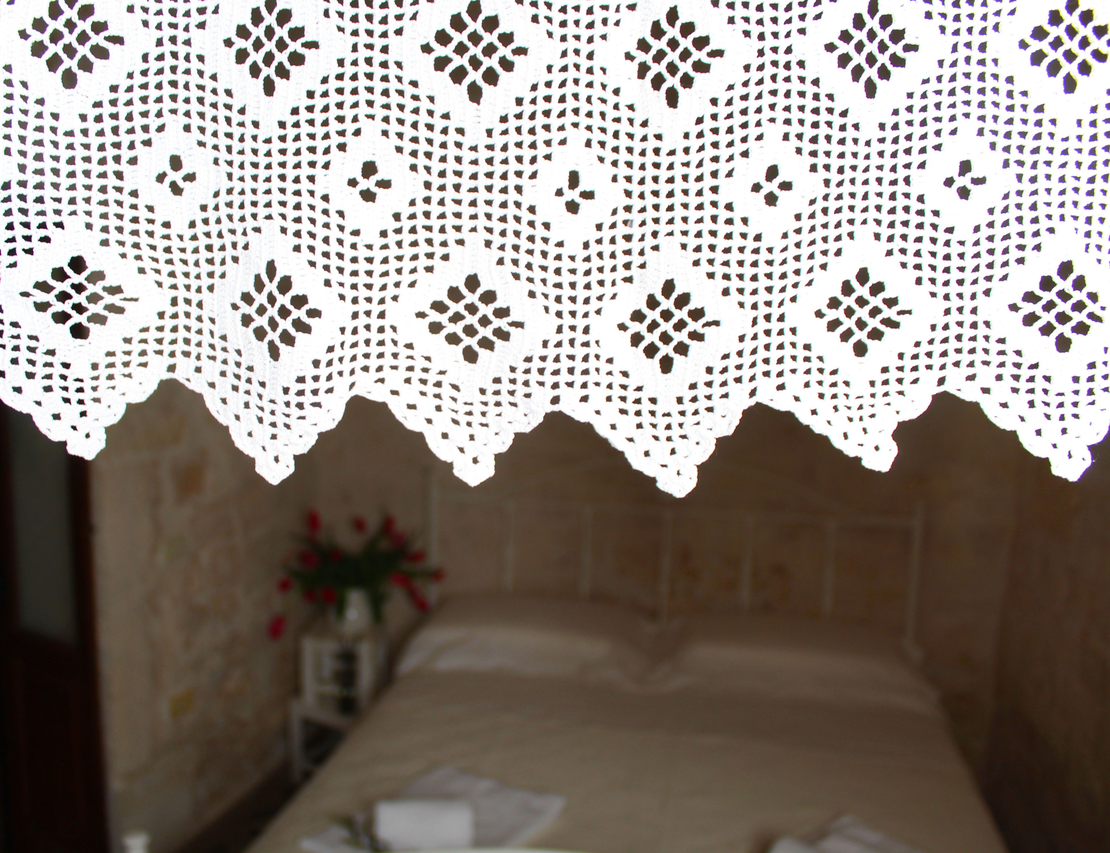 A room with a bed, a white lace headboard, and a vase with flowers on the side.
