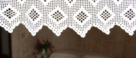 A room with a bed, a white lace headboard, and a vase with flowers on the side.