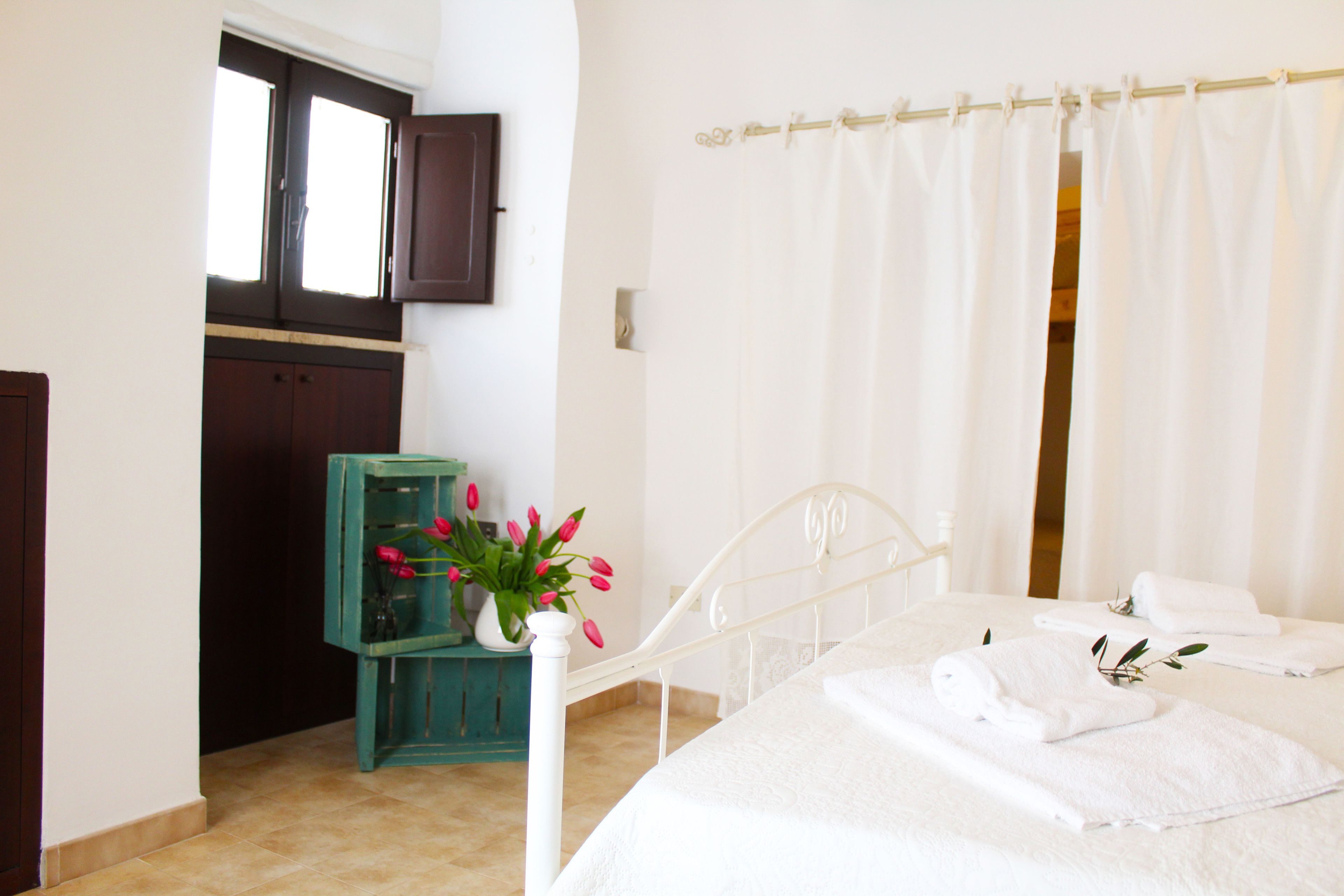 A bedroom with a bed, white curtains, a window, a door, and a small table with a vase of flowers.