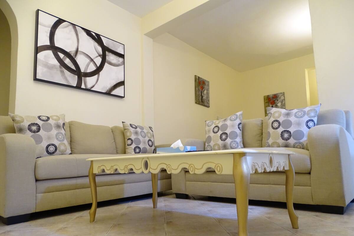 A living room with a beige sofa, patterned cushions, a white coffee table with gold legs, and abstract art on the wall.