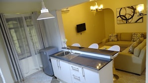 A compact kitchen with a gas stove, a sink, and a small refrigerator. There is a dining area with a table and chairs, and a sofa in the living room. - Nyali Parkside Resort (Mombasa)