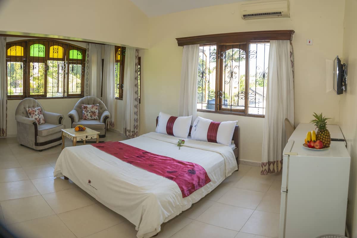 A bedroom with a bed, two armchairs, a small table with fruit, and a refrigerator.