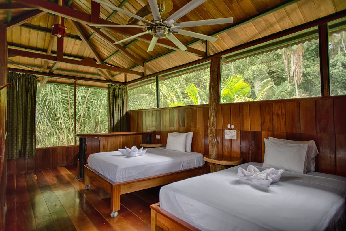 A room with two beds, wooden walls, a ceiling fan, and large windows with a view of greenery.