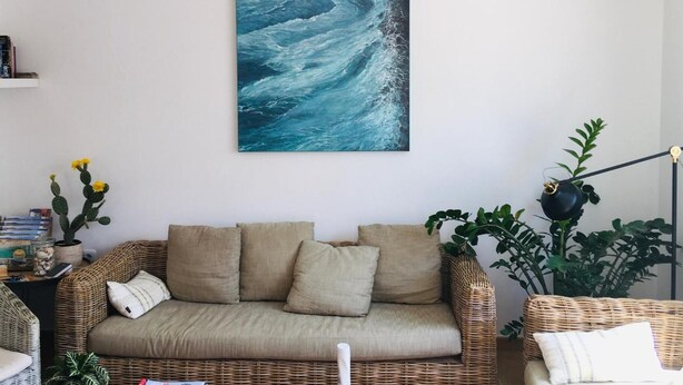 A living room with a wicker sofa, a painting of ocean waves, a hanging light fixture, and green plants.