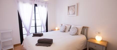 Un dormitorio ordenado con una cama grande, lámparas de noche, una mesita de noche y una ventana con cortinas.