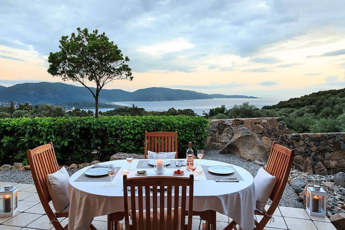 Un espace de restauration en plein air avec une table dressée pour quatre personnes, entouré de murs en pierre et offrant une vue sur la mer et les montagnes.
