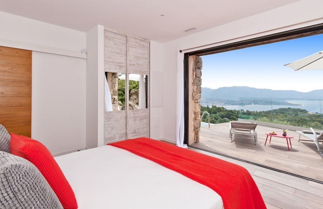 A bedroom with a large bed, a view of a balcony with outdoor furniture, and a stone wall.