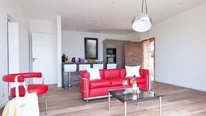 Een moderne woonkamer met een rode bank, een glazen salontafel en een rode fauteuil. In de achtergrond is een keuken zichtbaar met een wit aanrechtblad en een hangende lamp.
