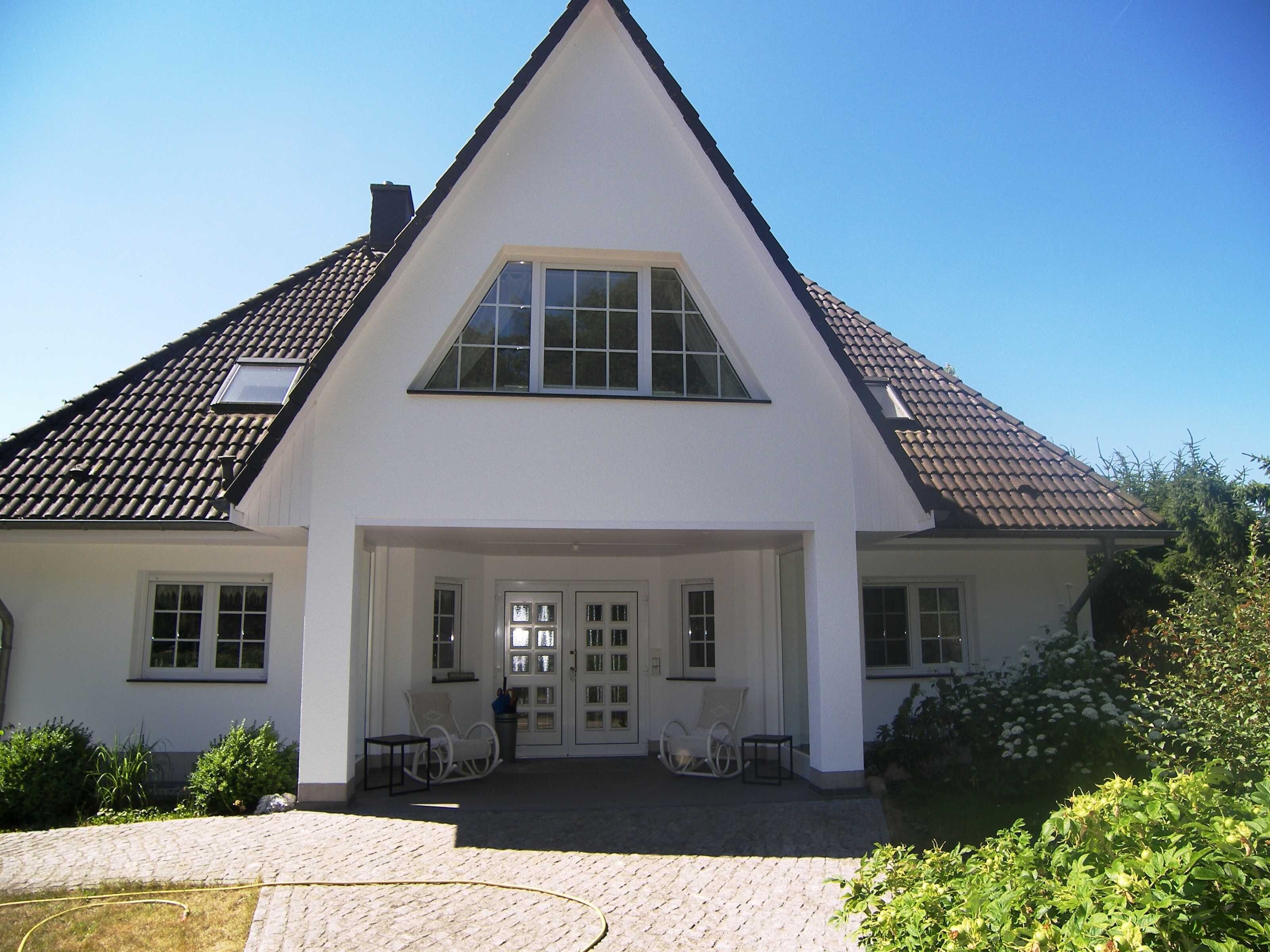 A large, white, two-story house with a steeply pitched roof, a front porch with seating, and a clear blue sky.