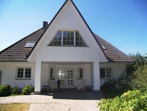 Een groot, wit huis met twee verdiepingen, een steil dak, een veranda met zitplaatsen en een helderblauwe lucht.