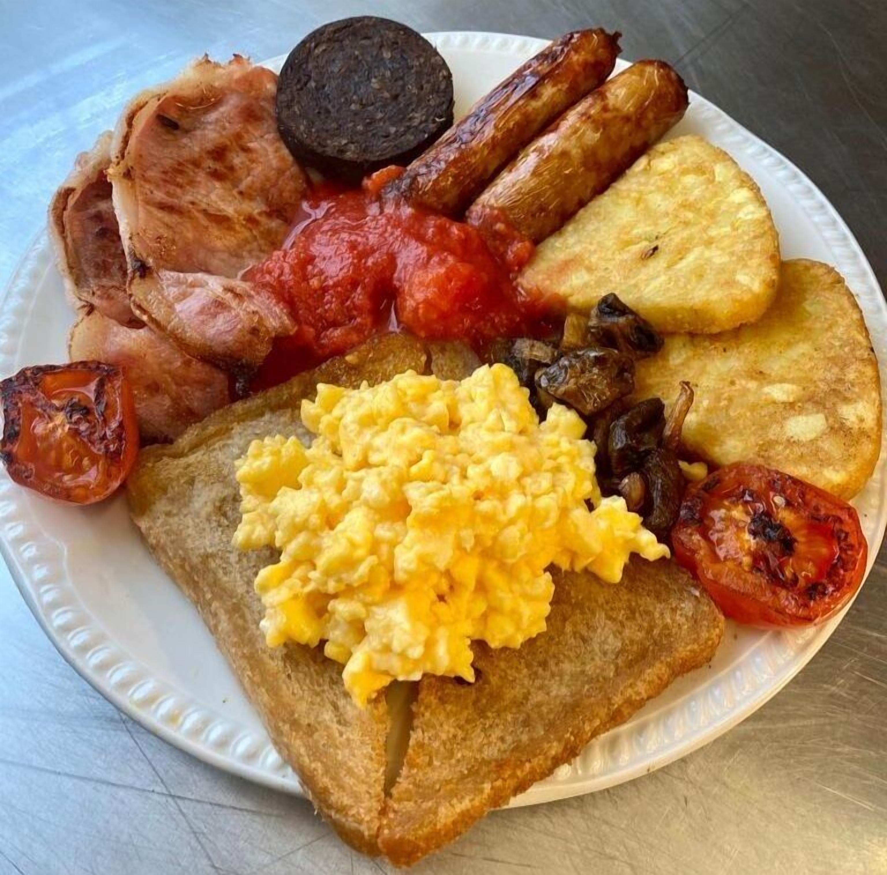 A plate of breakfast food including toast, scrambled eggs, sausages, bacon, tomatoes, and black pudding.