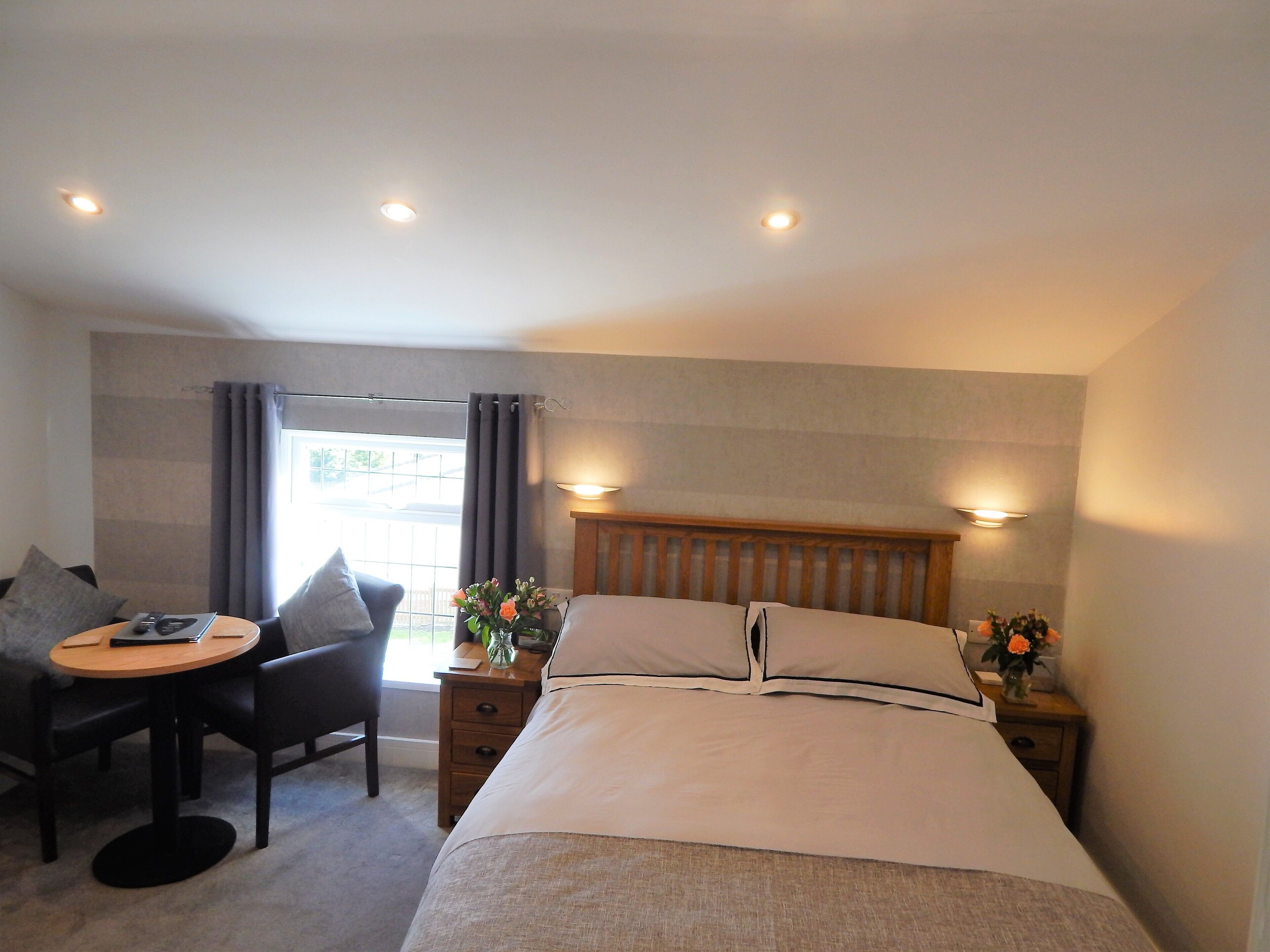 A hotel room with a bed, a desk, two chairs, a nightstand with a lamp, and a window with curtains.