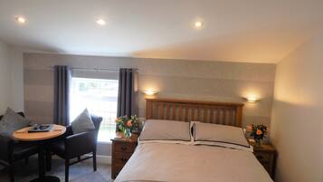 A hotel room with a bed, a desk, two chairs, a nightstand with a lamp, and a window with curtains.
