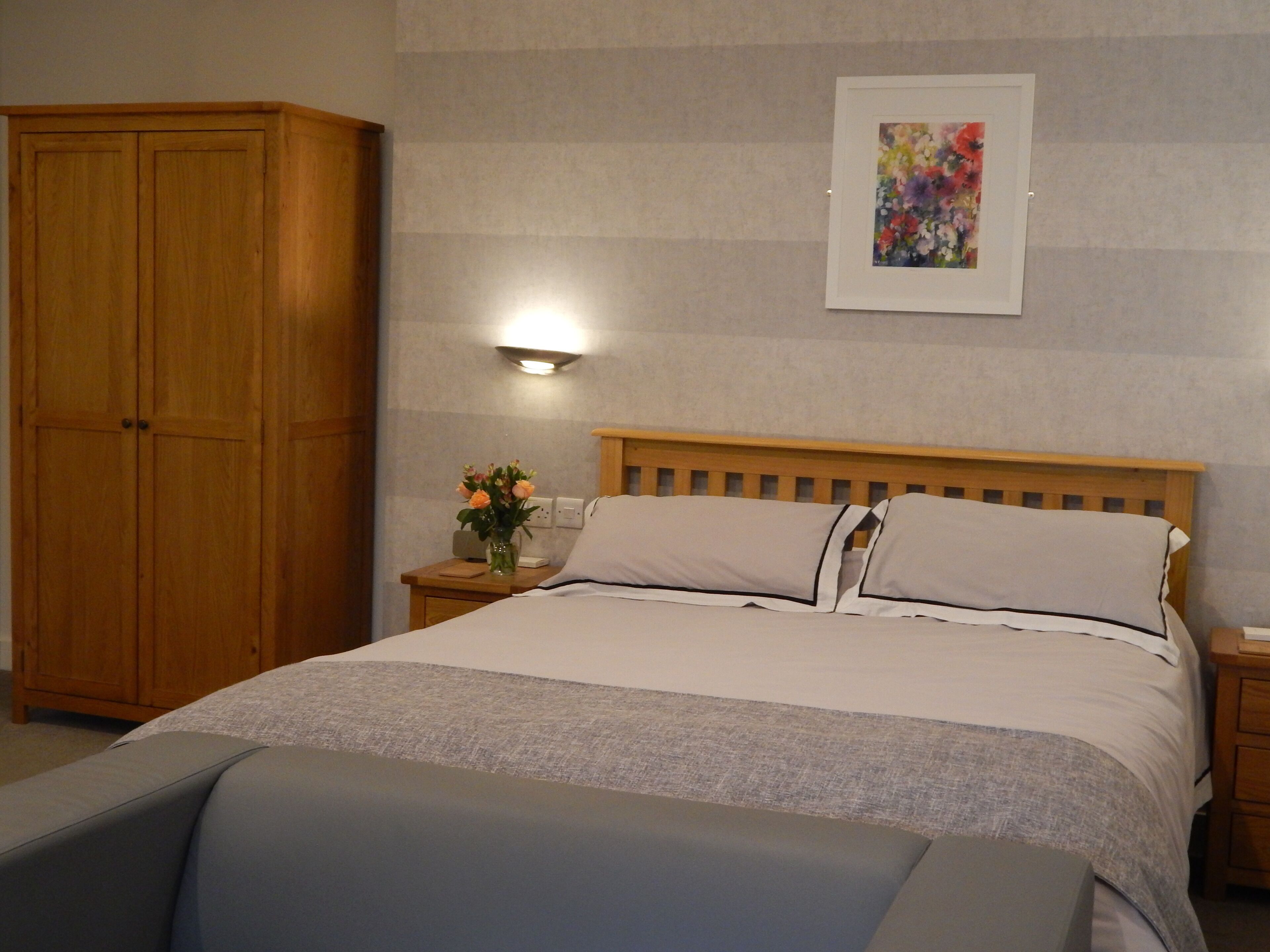 A bedroom with a wooden wardrobe, a bedside table with a vase of flowers, a bed with white bedding, and a framed artwork on the wall.