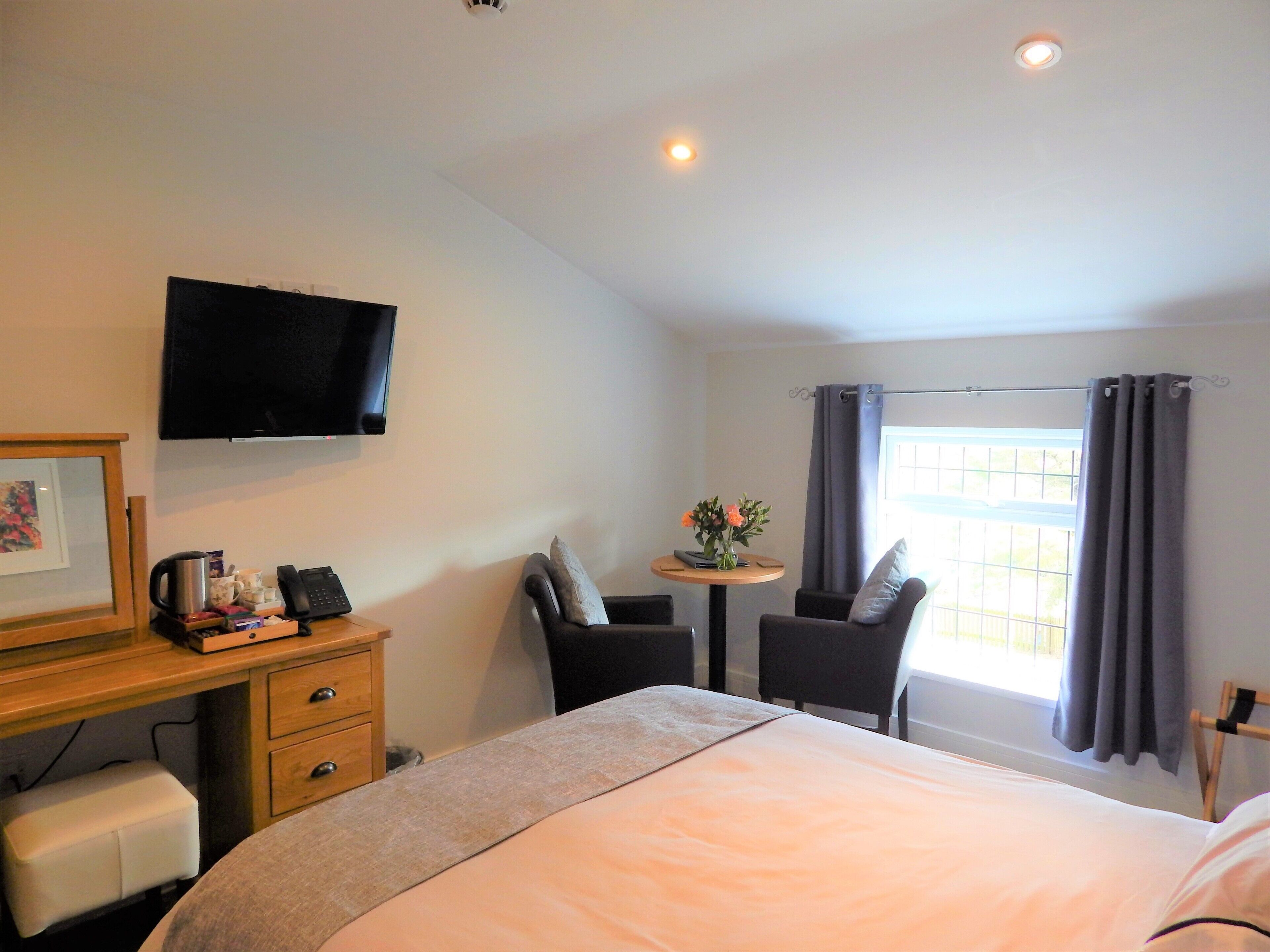 Ein Hotelzimmer mit einem Bett, einem Schreibtisch mit Computer, einem Sessel, einem kleinen Tisch mit Blumen und einem Fernseher an der Wand.